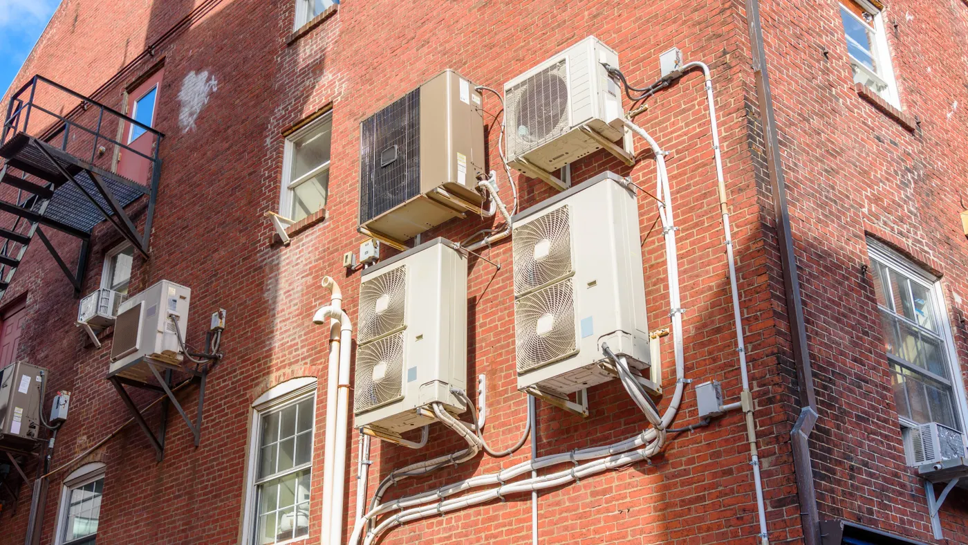 Heat pumps sit on the side of a brick building.
