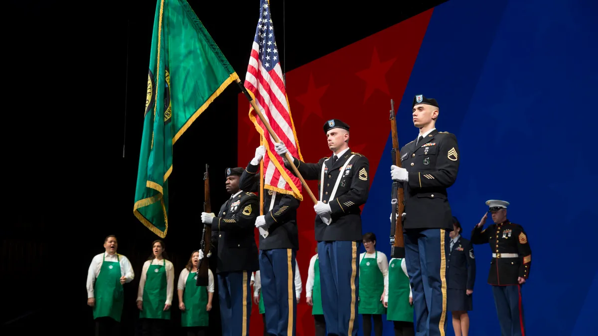 Starbucks workers in aprons sing, standing next to U.S. military color guards