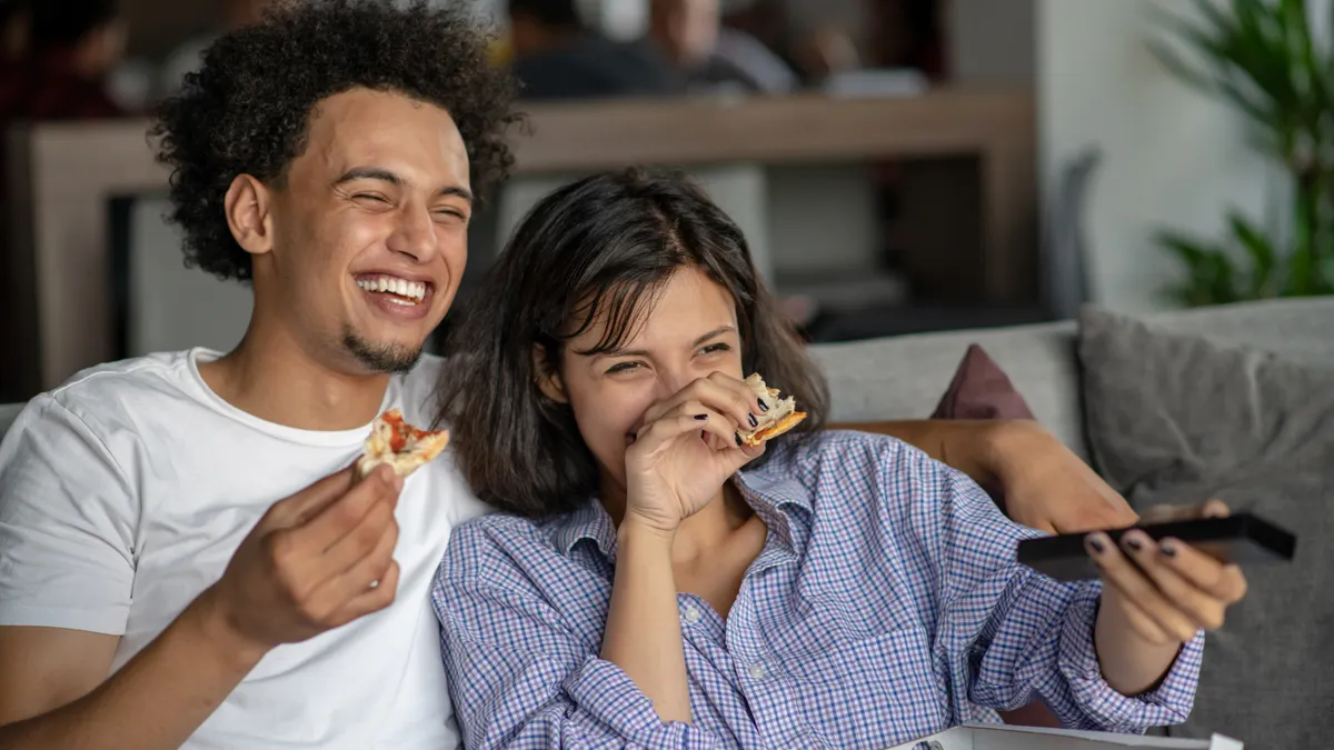 A joyful couple sits on a couch, sharing pizza and laughing while watching TV. The relaxed home setting conveys warmth and happiness.