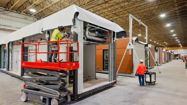 A worker on a scissor lift in personal safety equipment works on an exterior while another worker looks on.