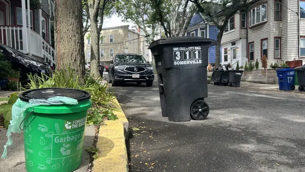 A small green bucket that says "Garbage to Garden" sits in the forefront of a street, with a large black bin and cars in the background.