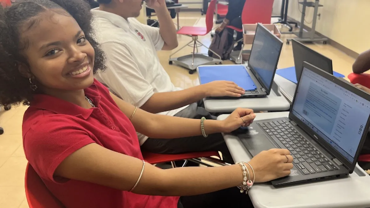 A student is sitting at a desk with an open laptop. They are looking at the camera and smiling.