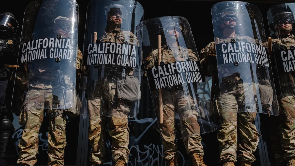 A line of people wearing camouflage suits and holding sticks and shields that say "California National Guard."