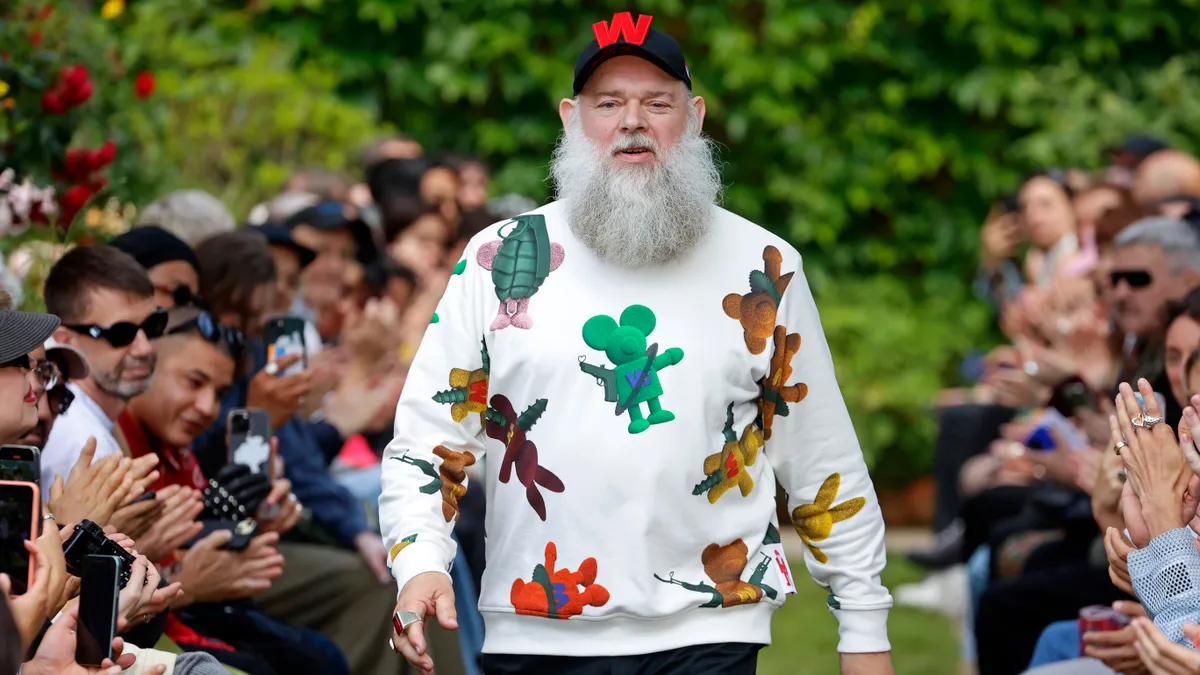 A person with a long white beard walks down a runway wearing a colorful top.