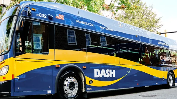 A blue and yellow bus lettered "100% Electric" and "DASH" on a street with trees.