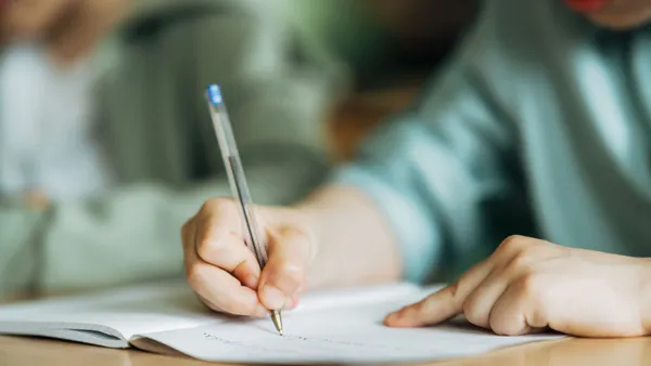A close-up of an elementary student writing with a pen in a workbook on a desk while studying in a classroom.