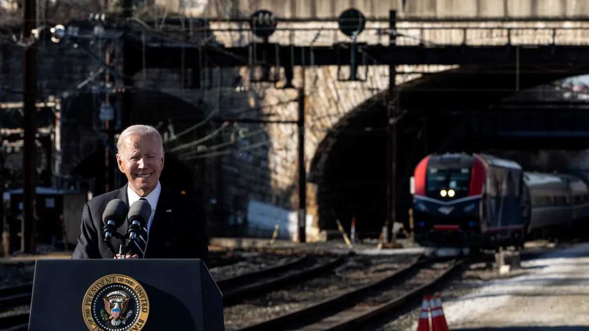 President Joe Biden, dressed in a dark suit with white hair, stands behind a podium in front of a brick tunnel and train.