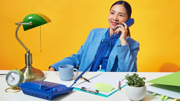 A woman in a blue suit is smiling while talking on a blue corded landline phone in a brightly colored office setup.