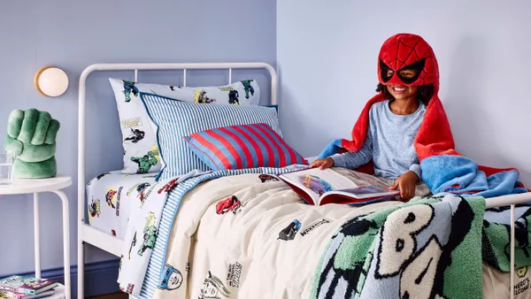 A child sitting on a Marvel-themed bed wearing a blanket with a Spiderman mask.