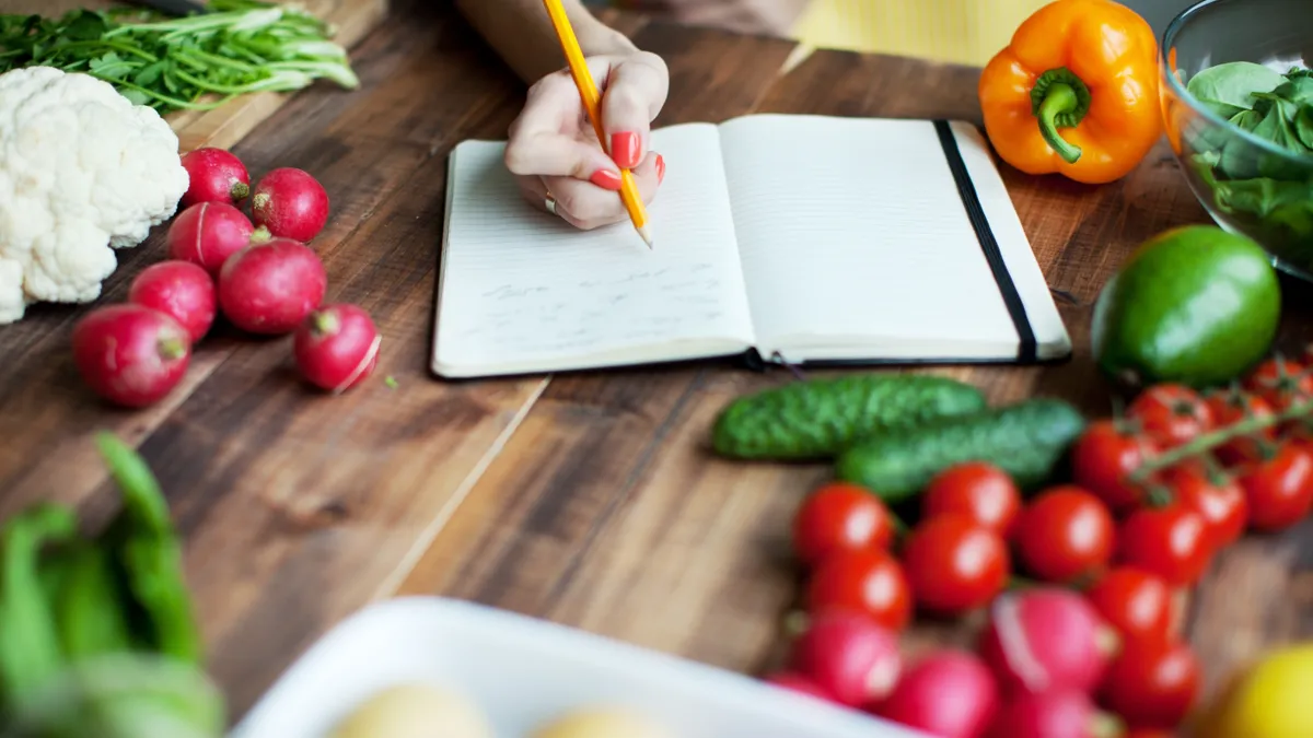 A wood counter with fresh vegetables in front of a open notebook and a hand writing in it.