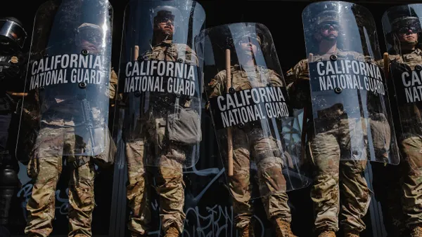 A line of people wearing camouflage suits and holding sticks and shields that say "California National Guard."