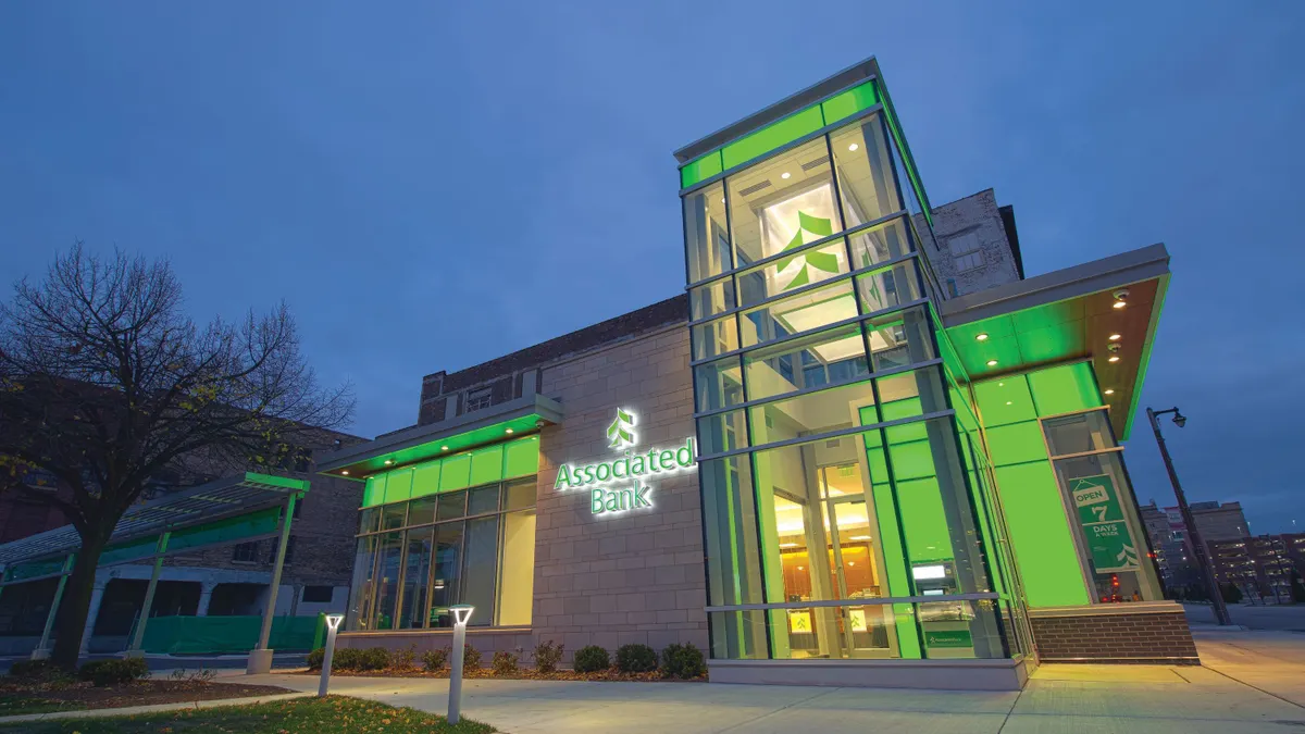 A glass and stone building is shown in the evening, with lights on inside, and "Associated Bank" name and logo on the outside.