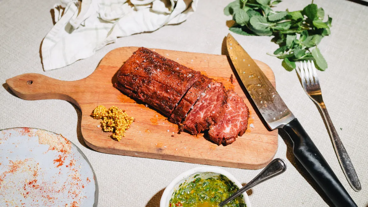 A partially sliced Juicy Marbles Plant-Based Loin Roast sits on a wooden cutting board on a textured white surface, A knife and mustard seeds are on the board, and a plate, sauce and herbs are near.
