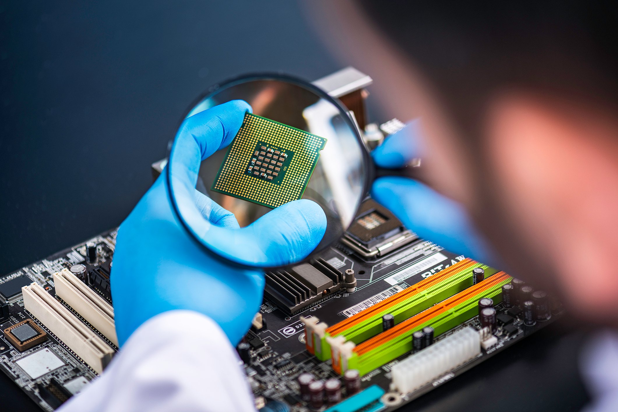 A person with blue latex gloves inspecting a gold and green computer chip with a magnifying glass.