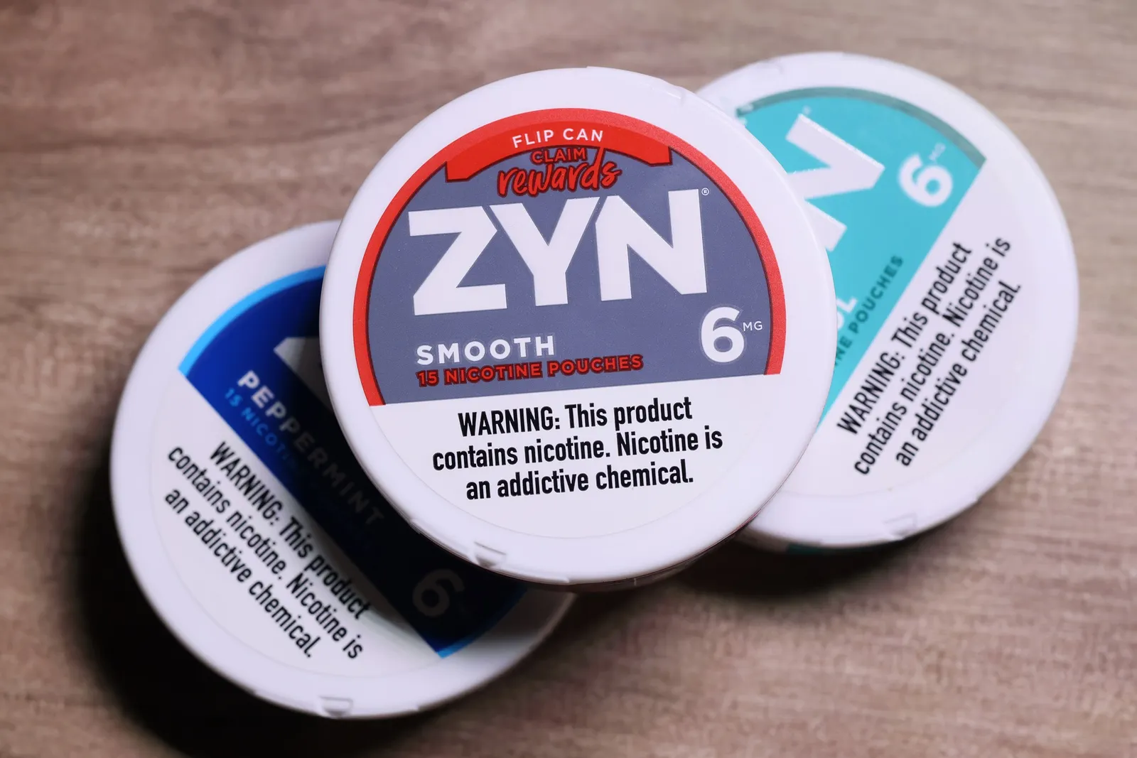 A photo of several containers of nicotine pouches. The top container says "Zyn smooth. Warning: This product contains nicotine. Nicotine is an addictive chemical."