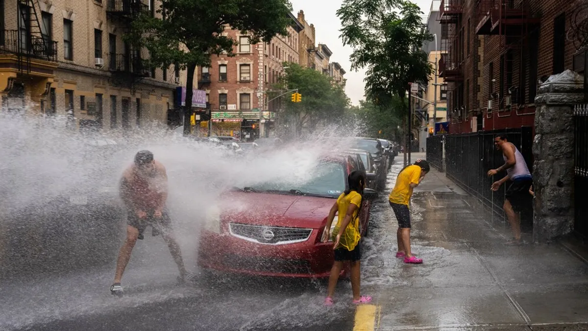 People get sprayed by water on a city street lined with cars.