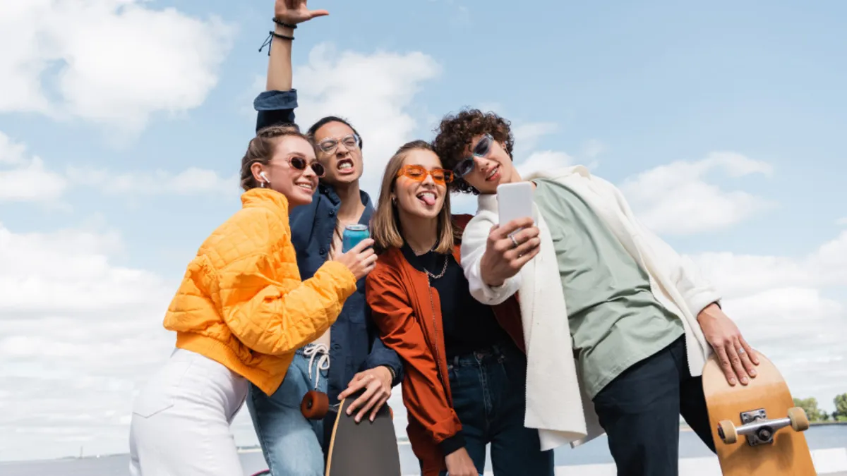 Four friends smiling and posing for a group selfie on a bright, sunny day, with two holding a skateboard.