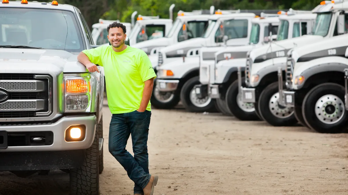 Man leaning on garbage truck in parking lot with other trucks