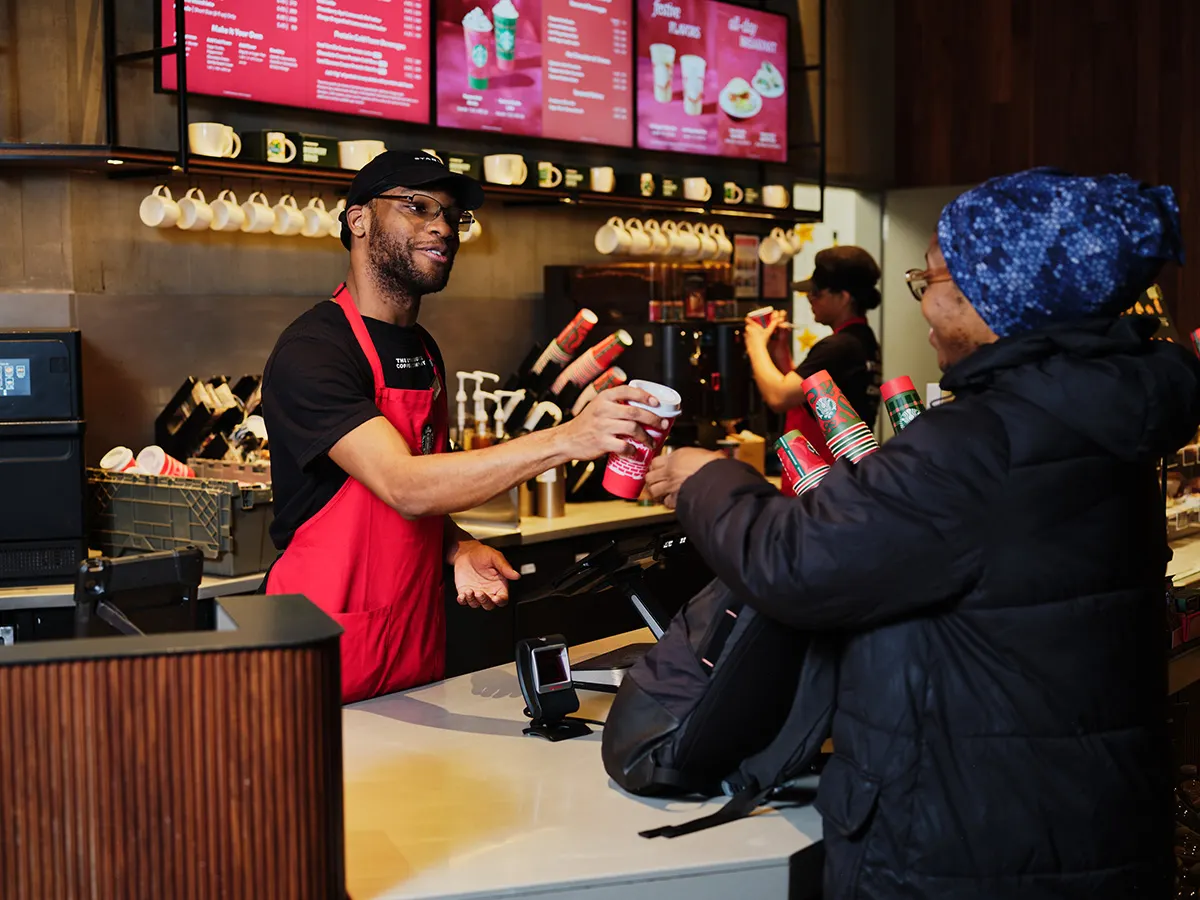 A person wearing a green apron hands off a reusable red cup to another person inside a coffee shop