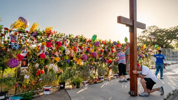 A person kneels at a large wooden cross as two people look at a fence covered with flowers and photos.