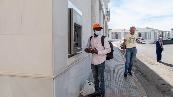 Person stands outside building in front of ATM with things in his hands and plastic bag at his feet, with two people walking by behind.