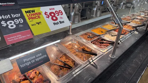 Hot prepared food items in a grocery store case with a sign that says "new lower price" at the top left.