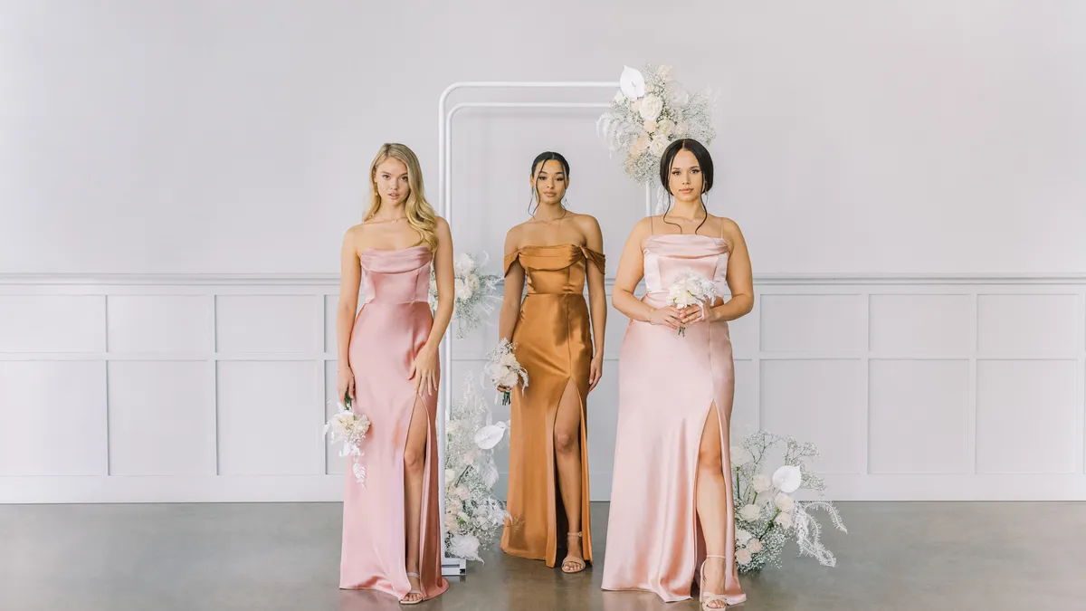 Three models in light pink and orange floor length dresses holding white flowers