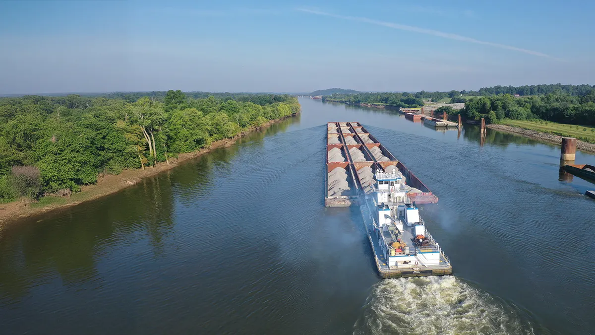 An aerial shot shows a tugboat pushing a barge on a river.