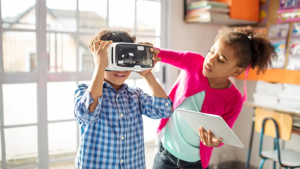 Girl holding digital tablet while assisting boy to wear VR headset. School children using technologies in classroom. They are in elementary school.