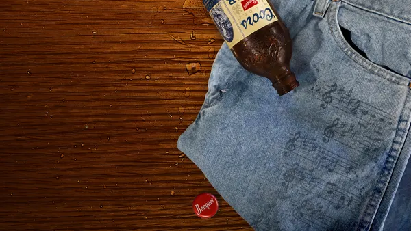 A pair of Wrangler jeans lay on a wooden table next to a glass bottle of Coors Banquet beer