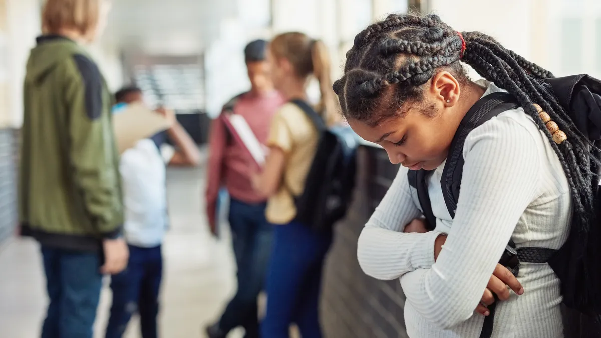 A young Black girl looks down, sadly, as she is excluded from peers in a school hallway.