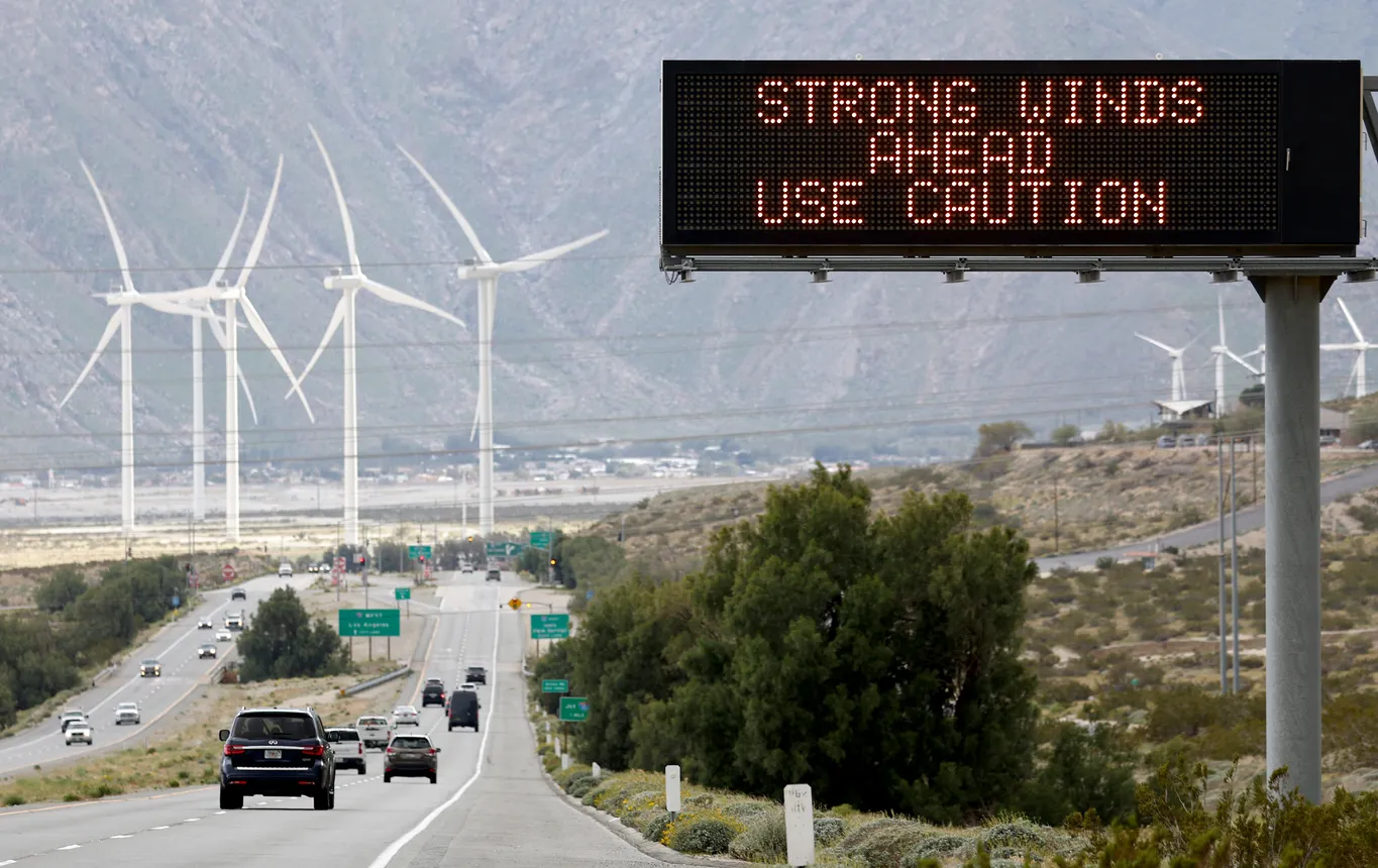Cars driving along highway with sign in foreground that says "Strong Winds Ahead, Use Caution" and wind Turbines in the background beyond the highway.