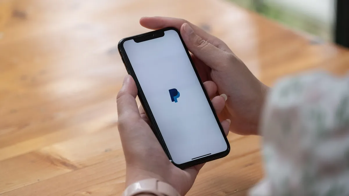 A person holds a phone at a table; the Paypal logo is on the screen.