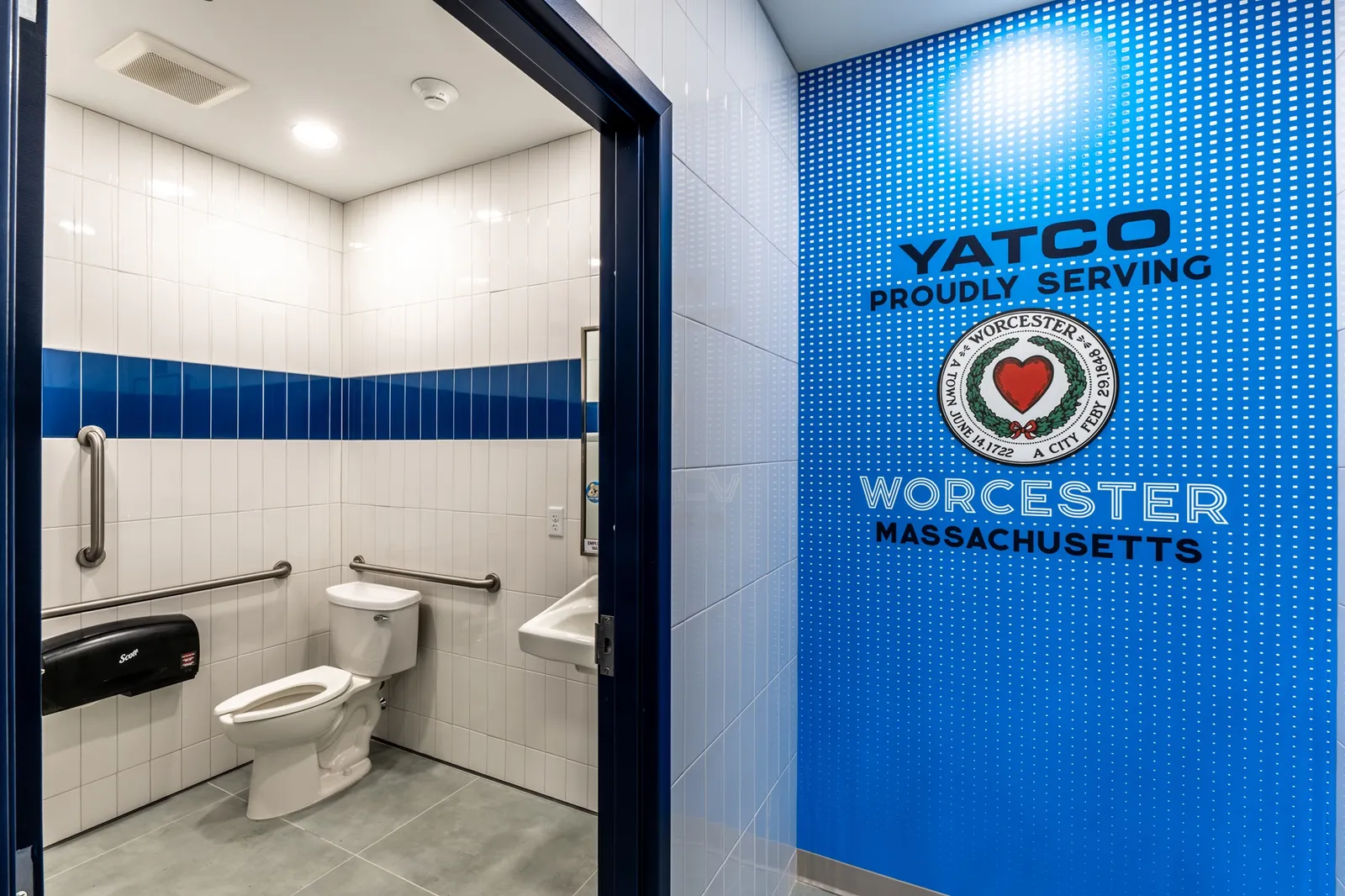 A photo of the interior of a store, showing a hallway and a bathroom through an open door. The bathroom has blue and white tile. A sign on the wall says Yatco. Proudly serving Worcester. Massachusetts.