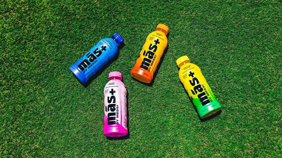 Four bottles of Mas+ by Messi on soccer turf.