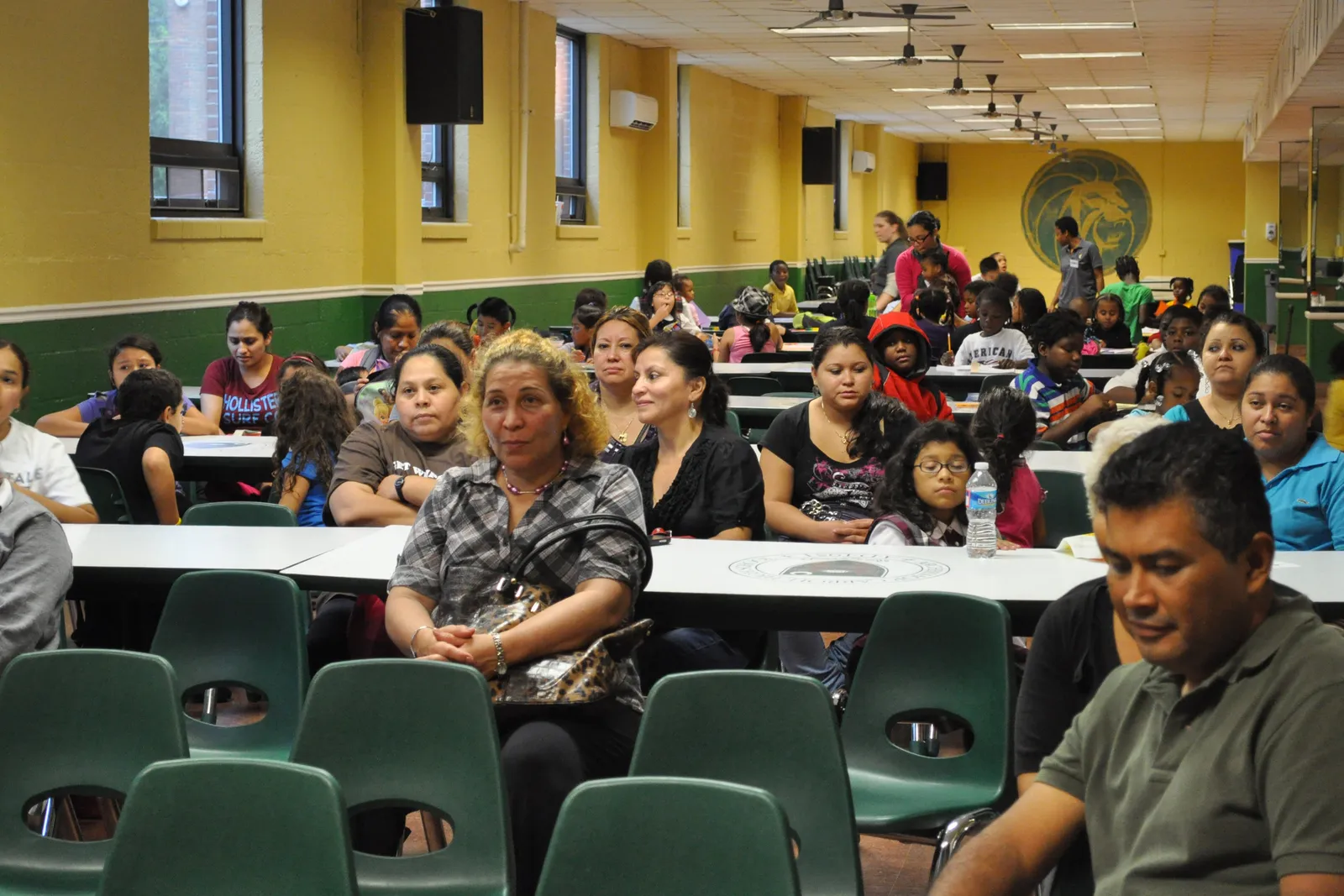 A roomful of people sit in chairs and at tables in a school cafeteria. They appear to be listening to a speaker.