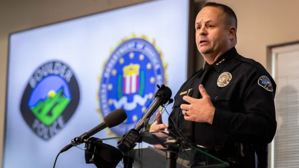 A person wearing a uniform with Boulder Police Department seal on left arm speaks into microphones in front of screen with a seal that says "Boulder Police."