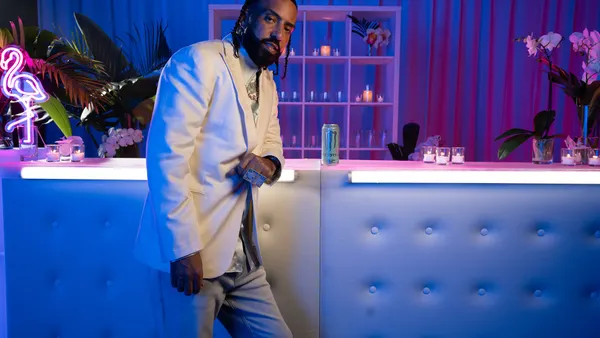 An image of a person standing in front of a bar, lit with candles and neon lights. The eprson is wearing a white jaclet, gray pants and a silver shirt. there is a can of energy drink on the bar.