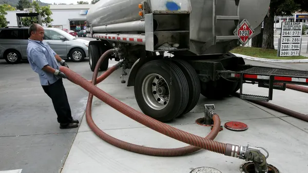 A photo of a person filling an underground gas tank from a truck. There is a gas station in the background.