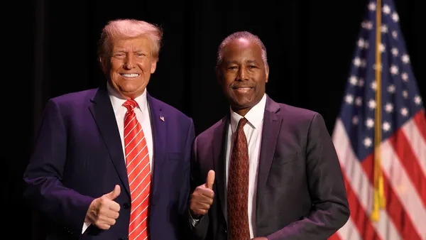 Two people wearing suits and ties pose for a photo, smiling and giving the thumbs up symbol. American flags are seen in the background.