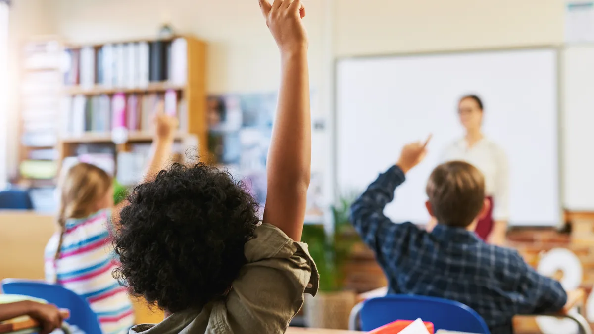 Young students sit at individual desks facing the front of the classroom with their hands raised.