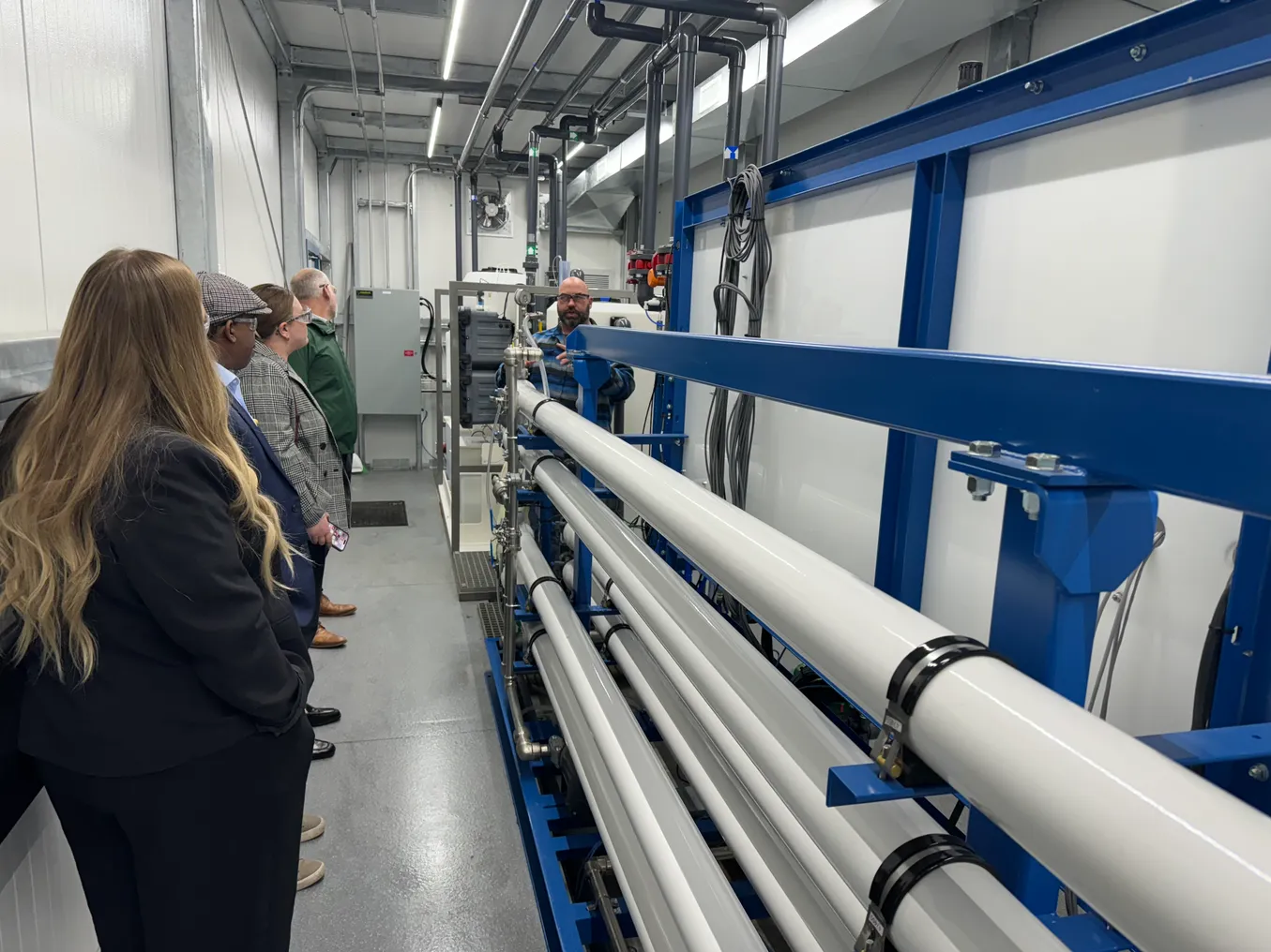 a group of people stand in a line in front of large white pipes in an indoor facility.