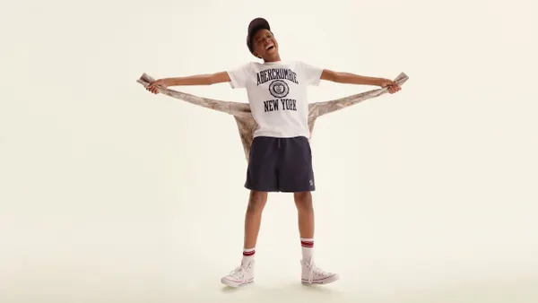 A smiling kid in a white printed t-shirt, dark shorts and white sneakers and socks.