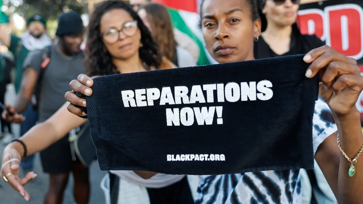 A protester displays a sign reading "Reparations Now!" in front of a crowd of other protesters.