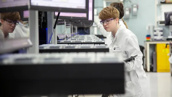 A person wearing goggles and holding a clipboard looks at a tray in a laboratory.