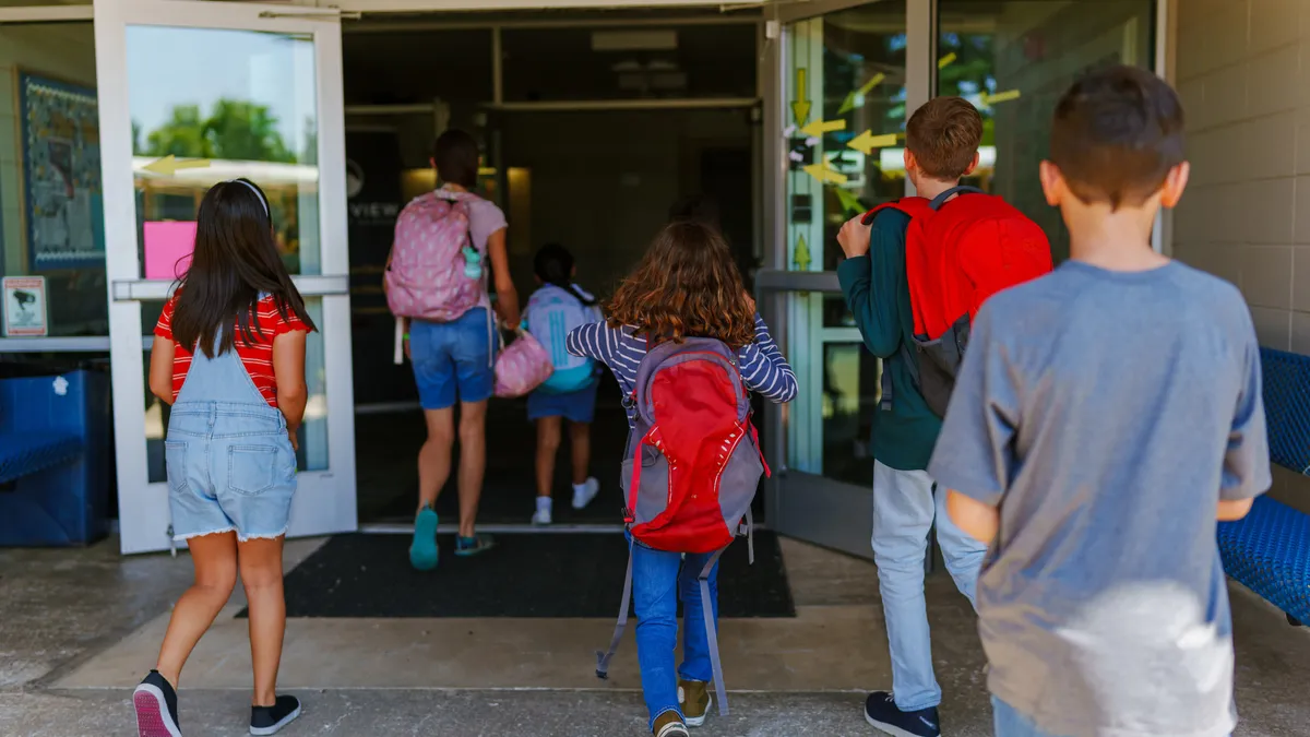 A group of elementary students enter a school building as they wear backpacks.