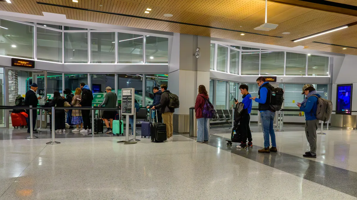 People stand in line to board a green bus seen through a glass partition.
