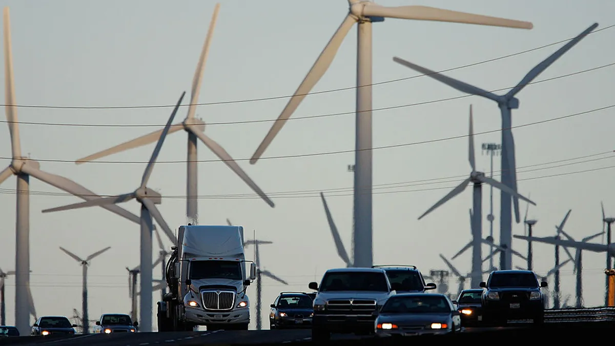 Diesel trucks and cars pass wind turbines along a freeway.