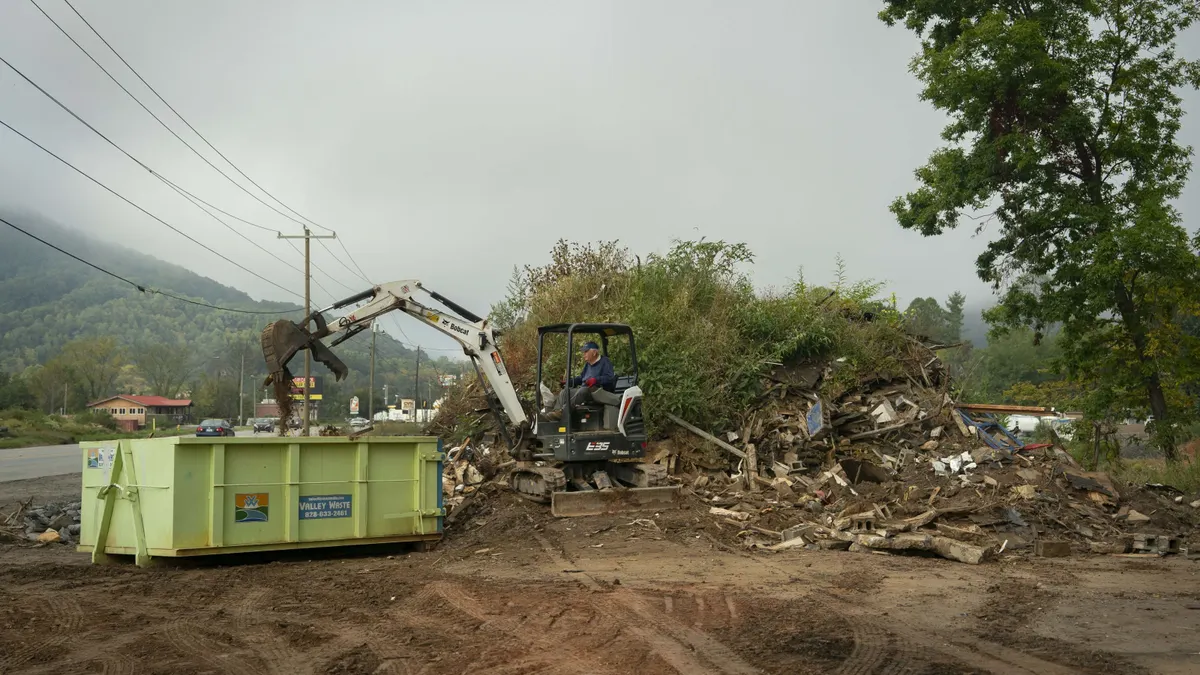 A man drives a small construction claw truck on a dirt lot near a road, transferring a large mound of wood waste and debris into a dumpster.
