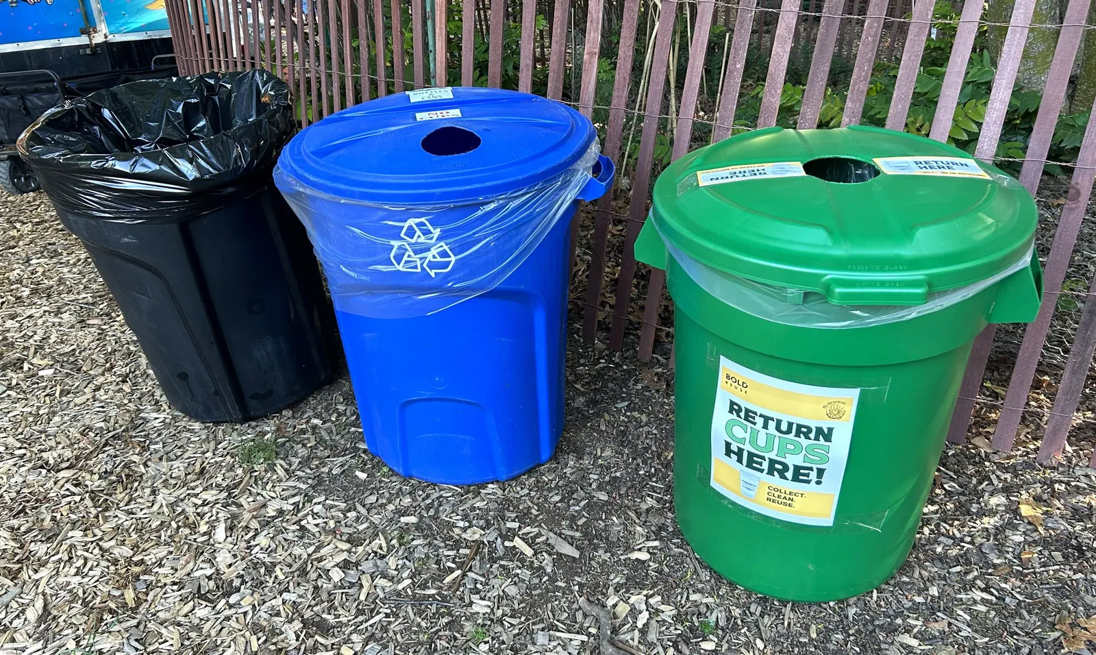 A black trash bin next to a blue recycling collection bin and a green reusable cups collection bin.